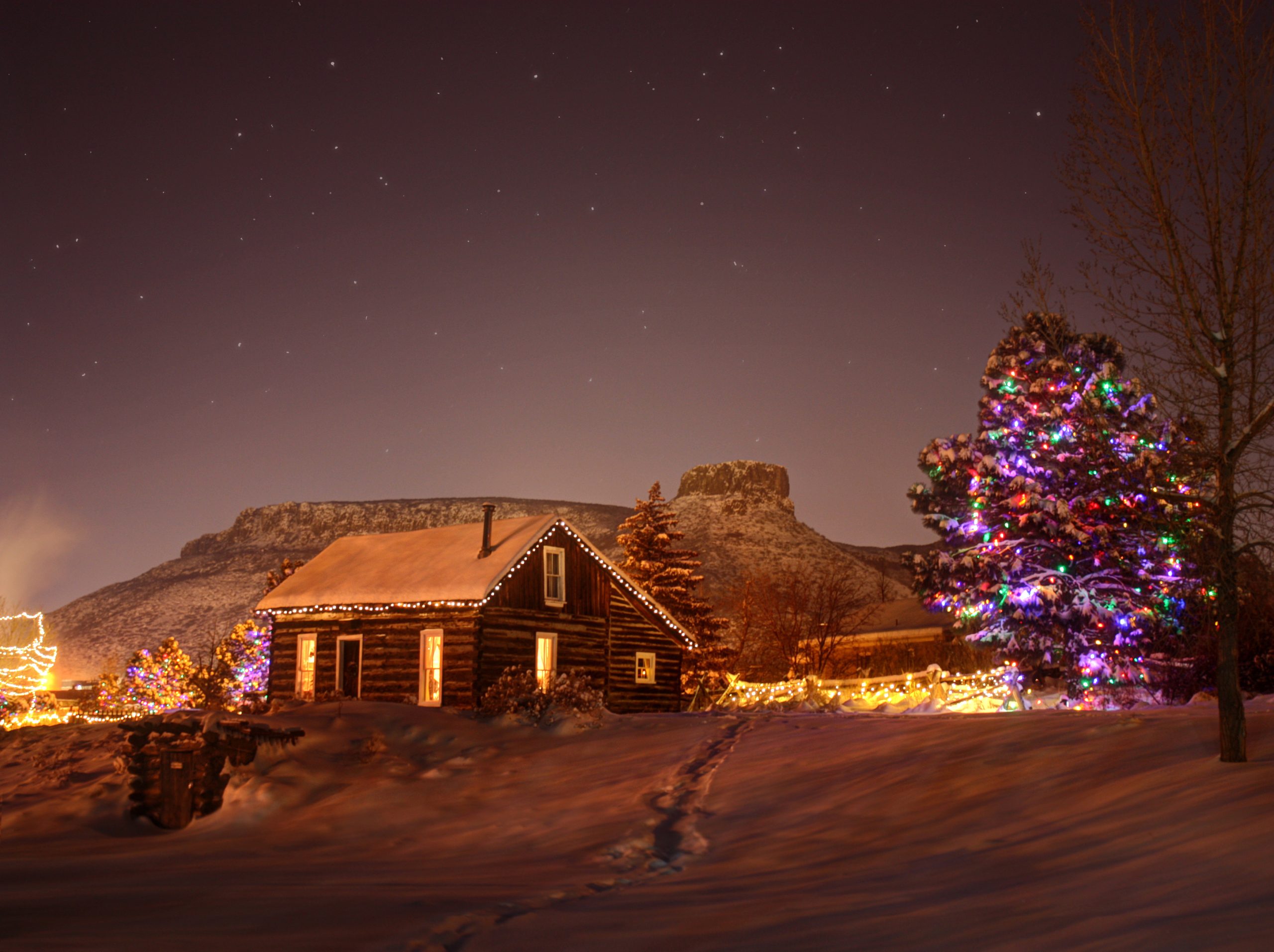 A snow covered Golden History Park at night with lights on inside the log cabin and trees in the park, castle rock is visible in the background