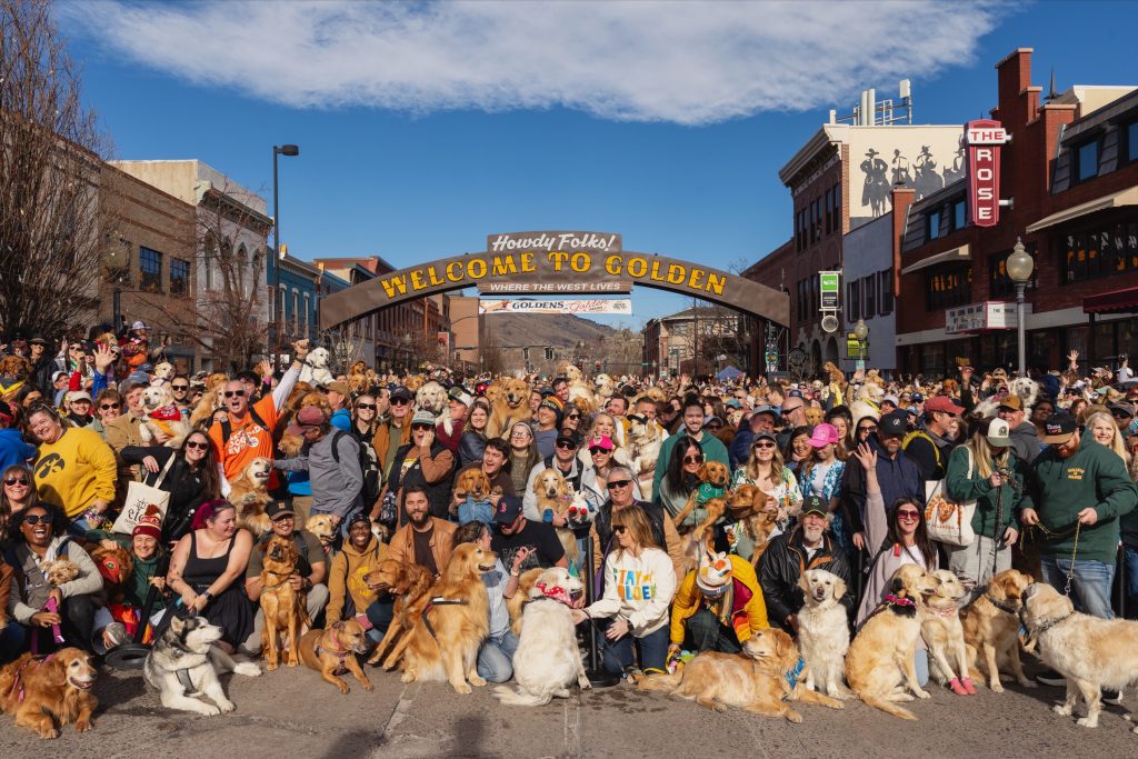 Goldens in Golden 2026 Group Photo of dogs and humans under the Welcome to Golden Arch