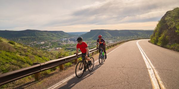 Two cyclers climbing Lookout Mountain on a winding road with a lush green landscape and a cityscape view in the distance under a sunny sky.