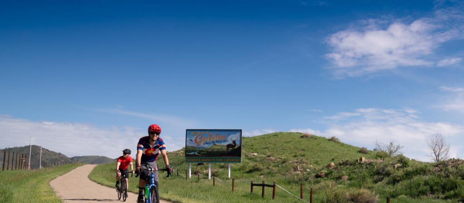 Two people biking on 6th Avenue paved trail past Welcome to Golden sign and Fossil Trace Golf Course