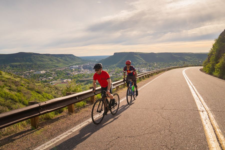 Two cyclers climbing Lookout Mountain on a winding road with a lush green landscape and a cityscape view in the distance under a sunny sky.