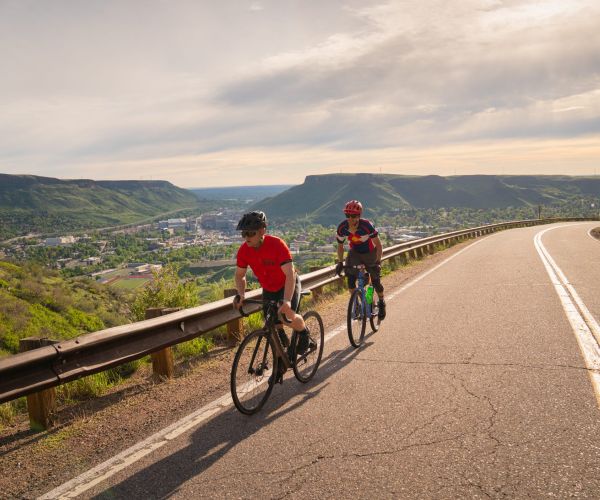 Two cyclers climbing Lookout Mountain on a winding road with a lush green landscape and a cityscape view in the distance under a sunny sky.